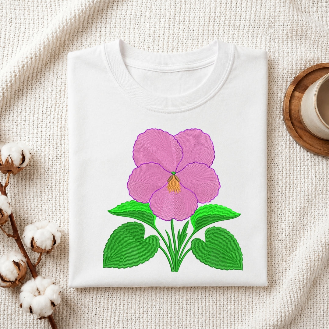 A white folded t-shirt featuring the embroidered pink pansy design, displayed on a textured white fabric with cotton sprigs and a wooden coaster.