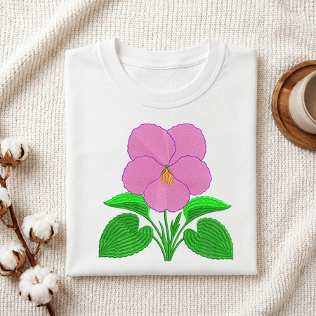 A white folded t-shirt featuring the embroidered pink pansy design, displayed on a textured white fabric with cotton sprigs and a wooden coaster.