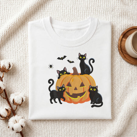 A neatly folded white t-shirt featuring the embroidered black cats and pumpkin Halloween graphic, displayed on a textured white knit background with cotton stems and a wooden cup.
