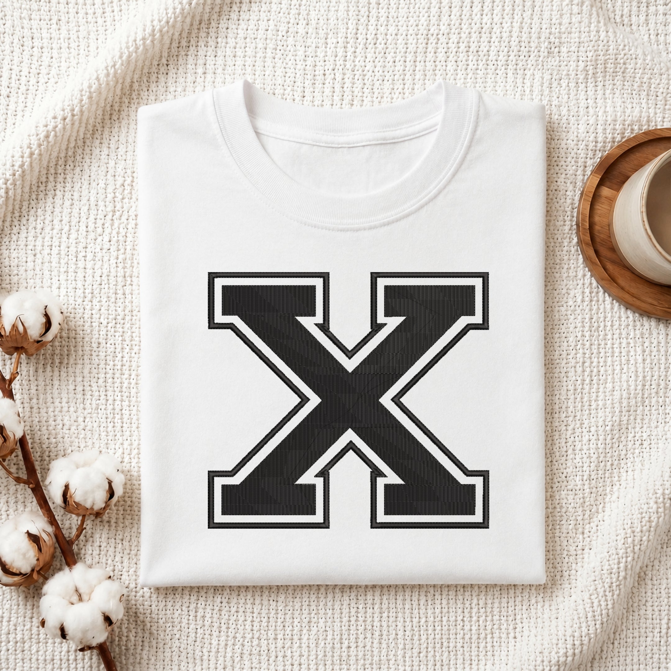 A folded white crewneck t-shirt with a large black and white letter 'X' graphic on the front, styled on a textured cream fabric with cotton stems and a cup on a wooden tray.