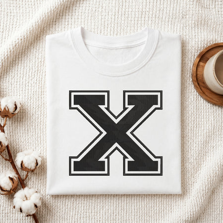 A folded white crewneck t-shirt with a large black and white letter 'X' graphic on the front, styled on a textured cream fabric with cotton stems and a cup on a wooden tray.