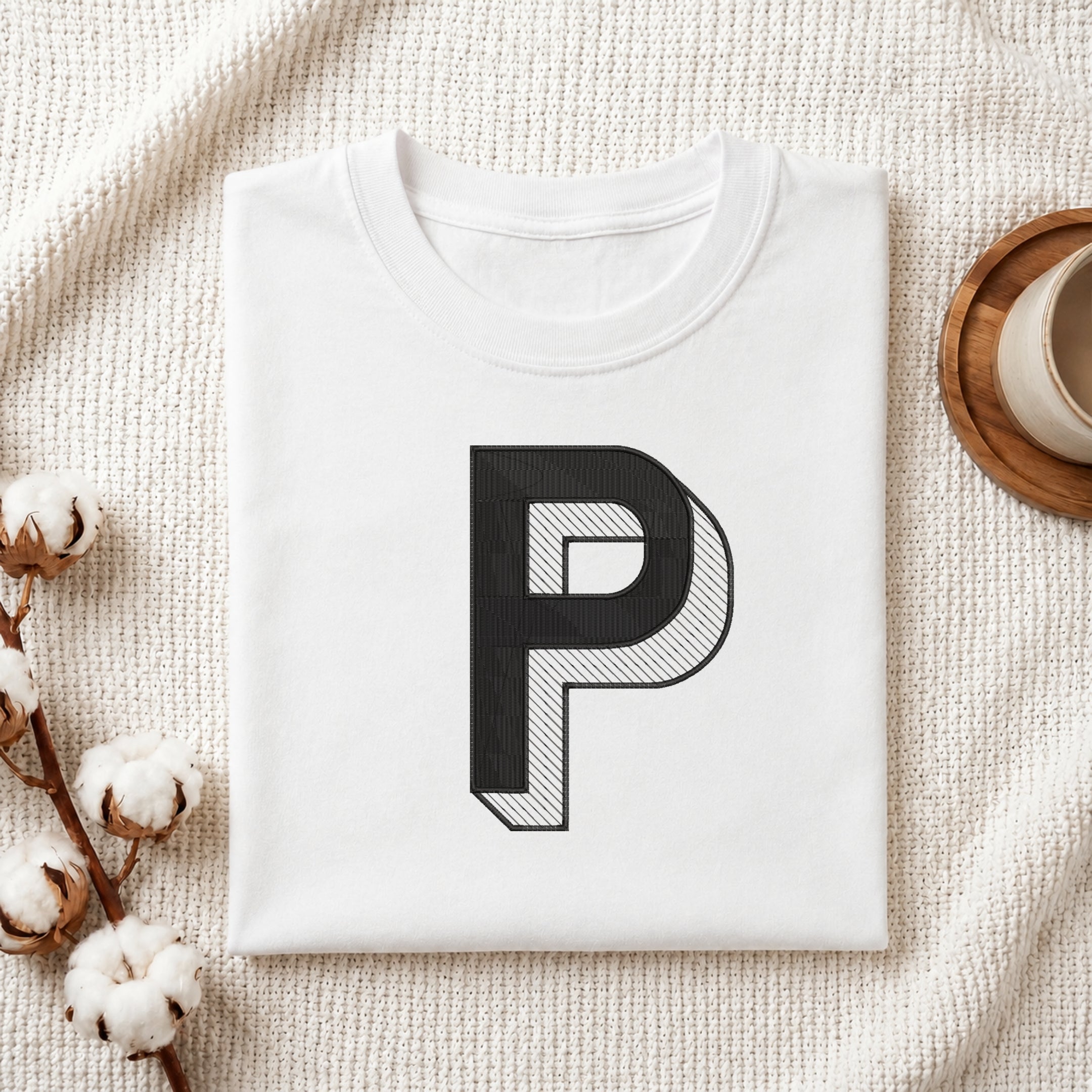 A folded white crewneck t-shirt with a large black letter 'P' graphic printed on the front, styled on a textured cream knit blanket with cotton bolls and a wooden cup in the background.