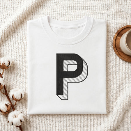 A folded white crewneck t-shirt with a large black letter 'P' graphic printed on the front, styled on a textured cream knit blanket with cotton bolls and a wooden cup in the background.