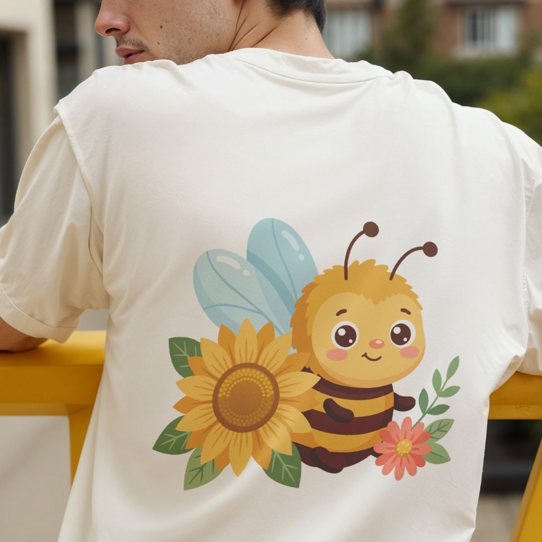 A man wearing a cream-colored t-shirt, viewed from the back. The shirt features a large graphic print of a cute cartoon bee with yellow and black stripes, a large yellow sunflower, a pink flower, and light blue wings.