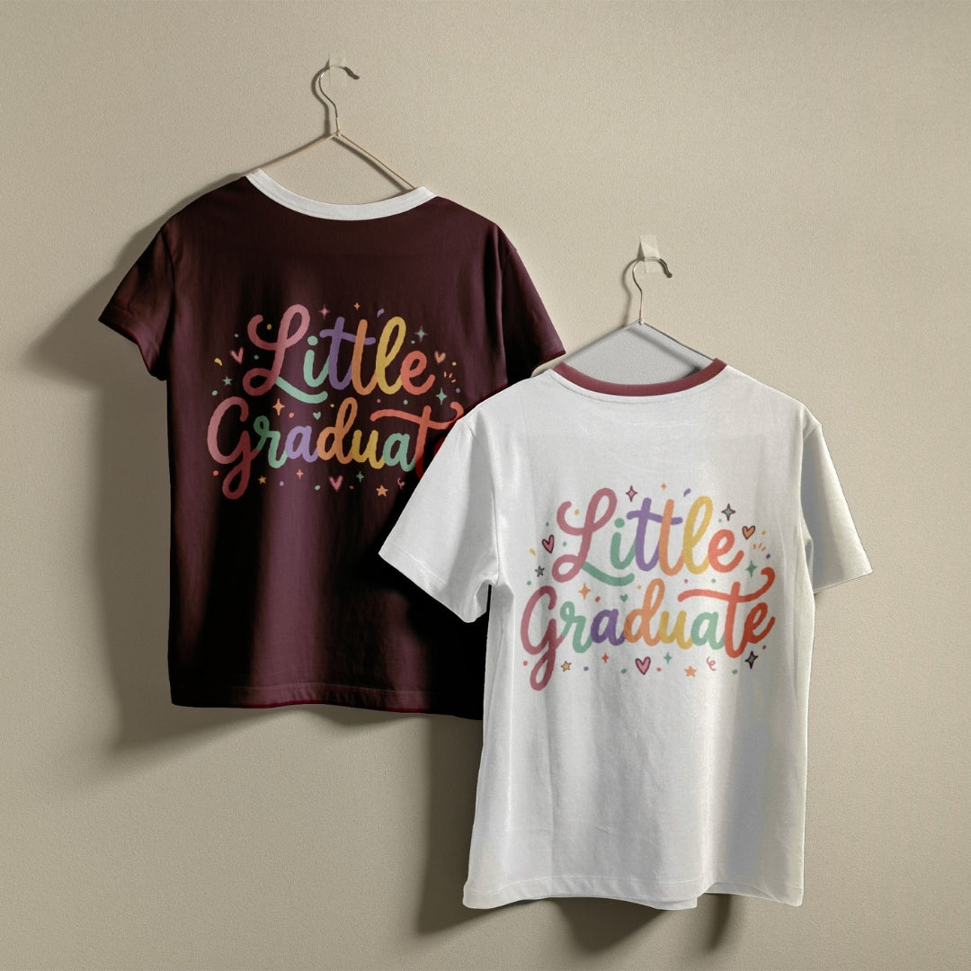 Two short-sleeve graphic T-shirts hanging on hangers against a plain beige wall. The back of one dark maroon shirt and the front of one white shirt both feature the words "Little Graduat" in a multicolor script with small star and heart accents.