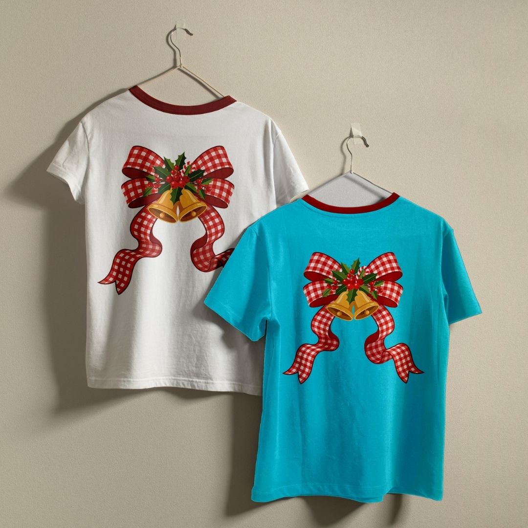Two children's short-sleeve t-shirts hanging on hangers against a plain beige wall. The left shirt is white with a red collar trim and a large red-and-white gingham bow with gold bells and green holly on the back.