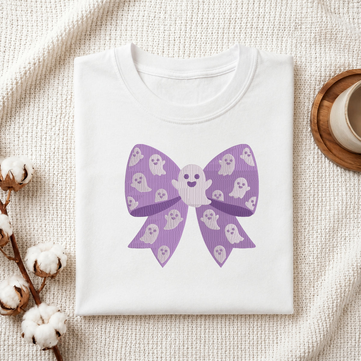 A neatly folded white t-shirt featuring the embroidered purple ghost bow design, displayed on a textured white knit background with cotton stems and a wooden cup.
