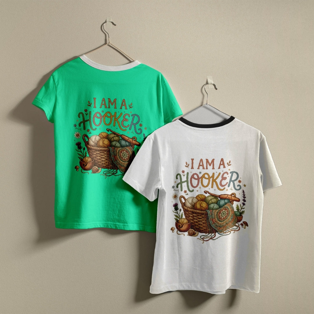 Two children's short-sleeve T-shirts hanging on hangers against a plain beige wall. The left shirt is bright green with a black collar trim and a colorful graphic on the back reading 'I AM A HOOKER' with baskets of eggs and autumn elements.