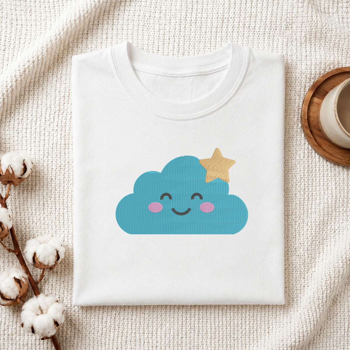 A folded white T-shirt with a cute blue cloud graphic and a gold star on the front, styled on a textured cream fabric with cotton stems and a small cup in the background.