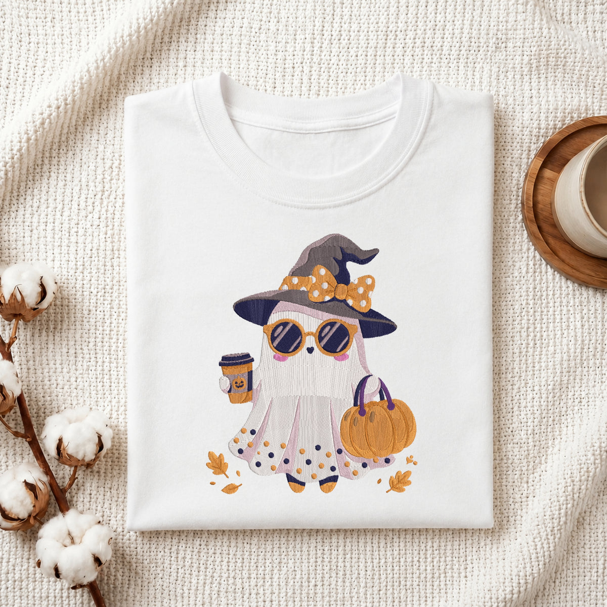 A folded white t-shirt with a cute cartoon ghost wearing a witch hat, sunglasses, and holding a pumpkin, styled on a textured cream fabric with cotton stems and a coffee cup prop.