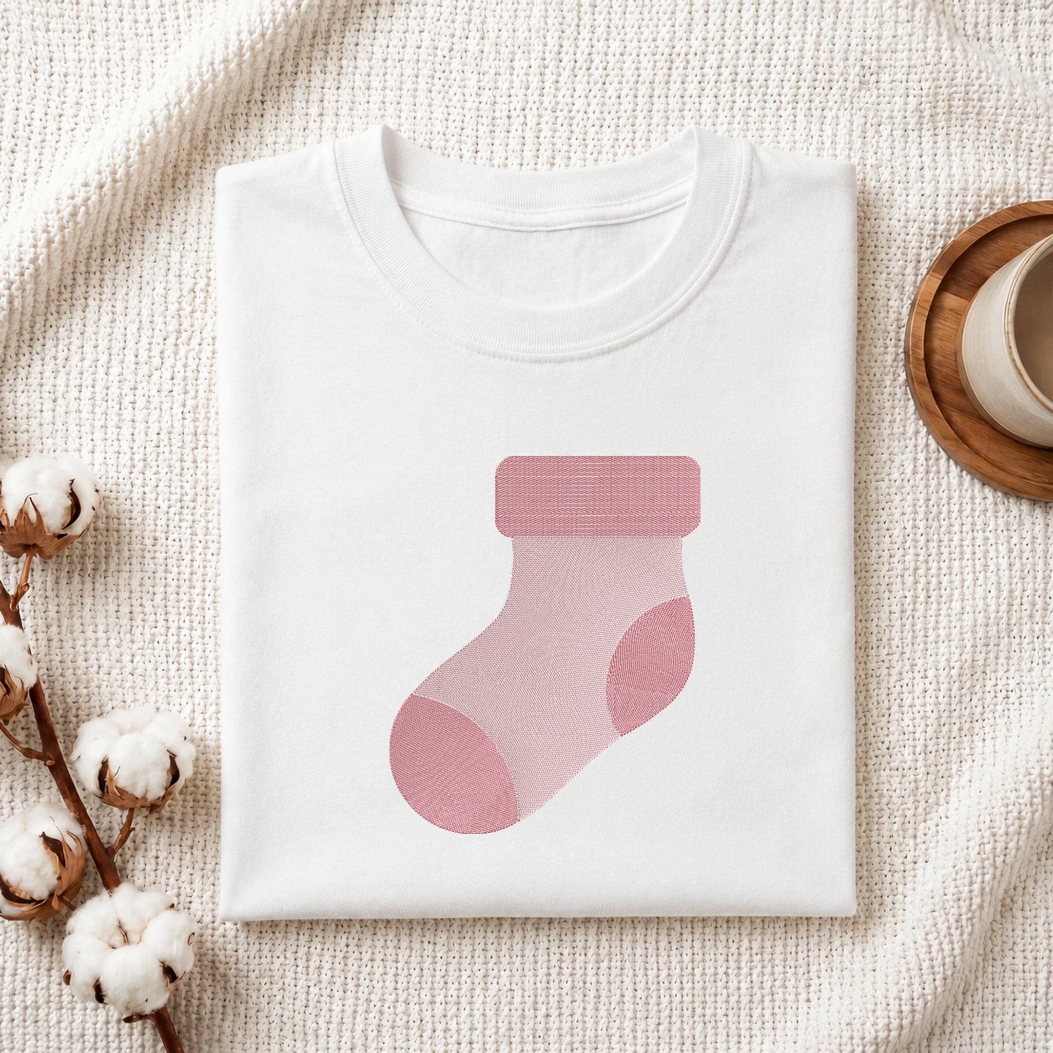 A folded white crewneck t-shirt featuring a pink sock graphic on the front, styled on a textured cream knit blanket with cotton bolls and a wooden cup in the background.