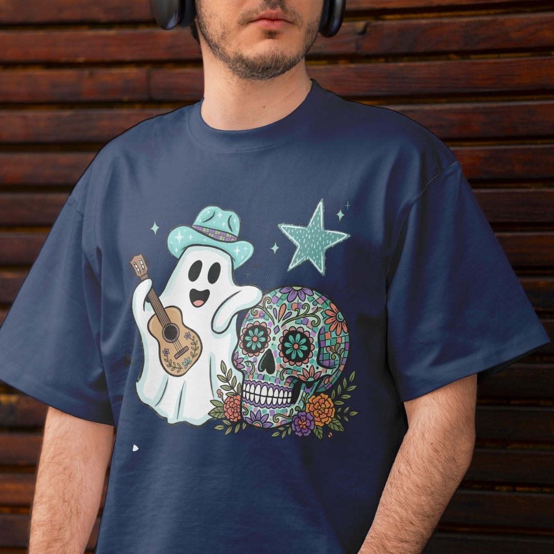 A man wearing a navy blue t-shirt with a colorful graphic of a ghost playing a ukulele next to a decorated sugar skull and a star, set against a dark wooden background.