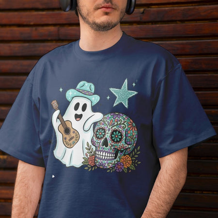 A man wearing a navy blue t-shirt with a colorful graphic of a ghost playing a ukulele next to a decorated sugar skull and a star, set against a dark wooden background.