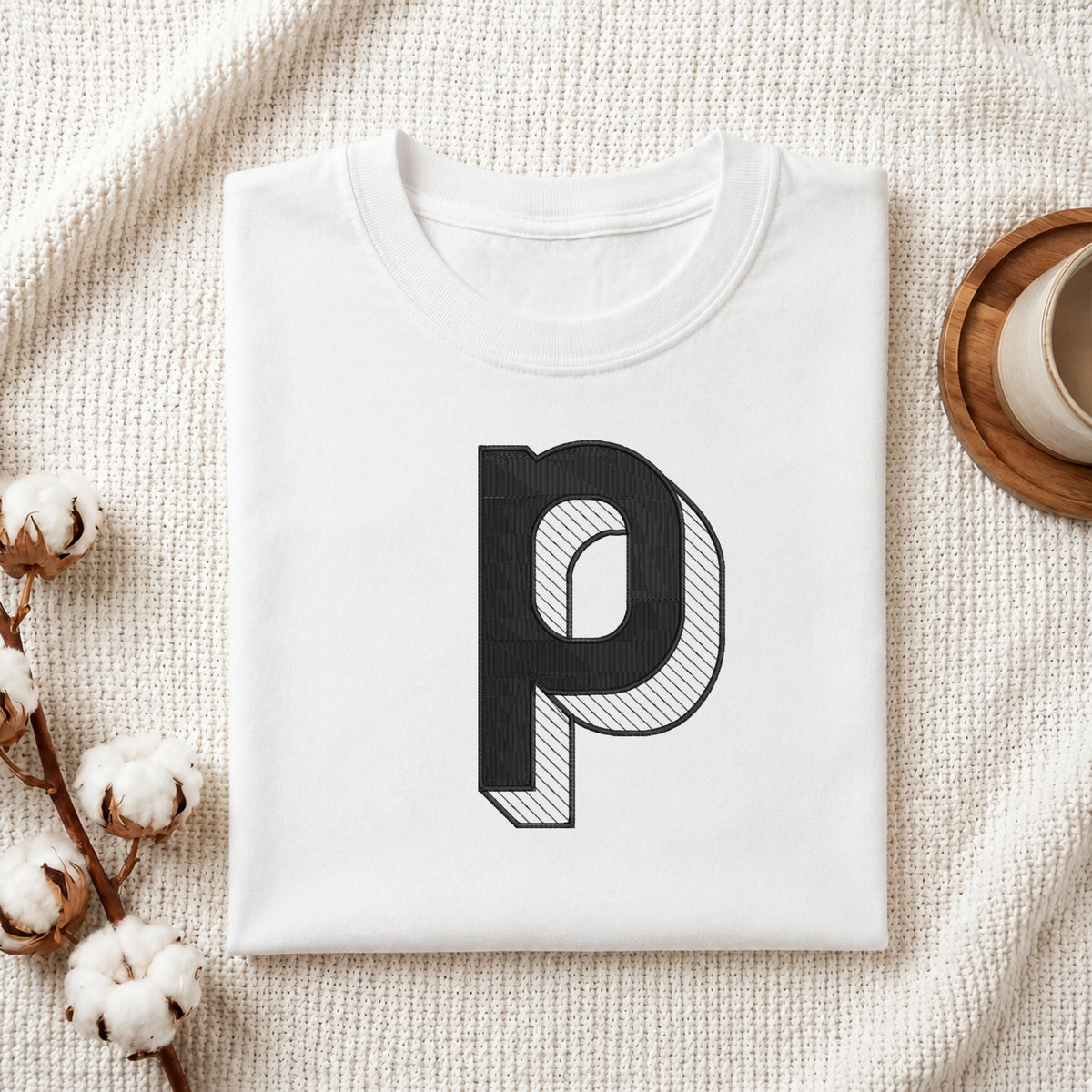 A folded white t-shirt with a large black letter 'p' graphic printed on the front, styled on a textured cream knit blanket with cotton bolls and a wooden cup in the background.