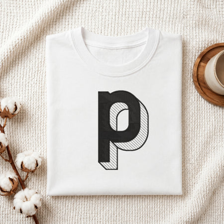 A folded white t-shirt with a large black letter 'p' graphic printed on the front, styled on a textured cream knit blanket with cotton bolls and a wooden cup in the background.