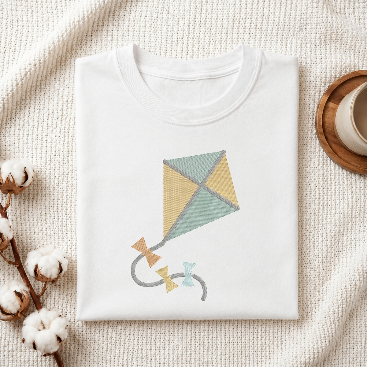 A neatly folded white crewneck t-shirt featuring a stitched cute kite design, styled with cotton branches and a wooden coaster.