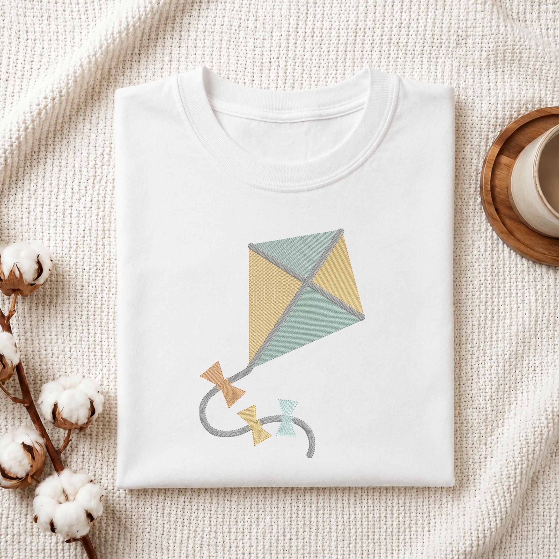 A neatly folded white crewneck t-shirt featuring a stitched cute kite design, styled with cotton branches and a wooden coaster.