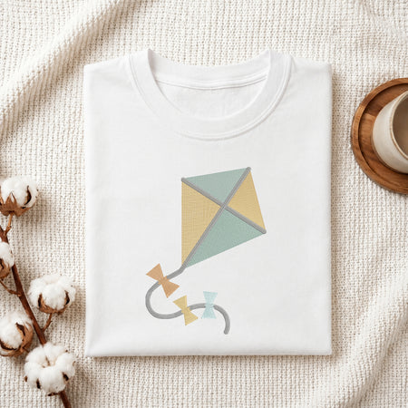 A neatly folded white crewneck t-shirt featuring a stitched cute kite design, styled with cotton branches and a wooden coaster.