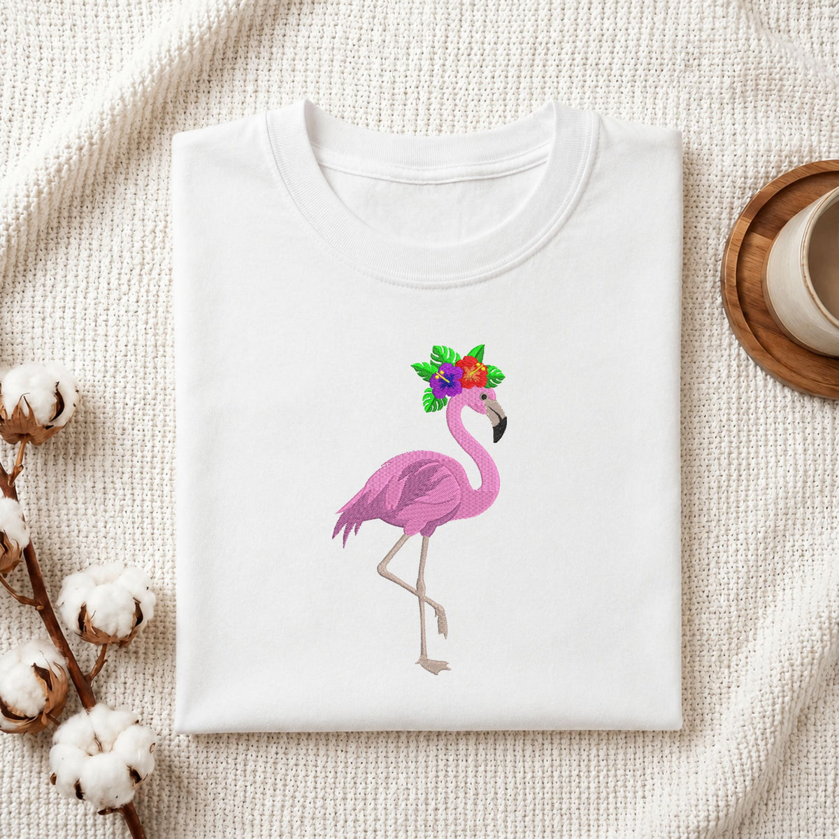 Folded white cotton t-shirt mockup featuring a tropical pink flamingo with floral crown machine embroidery design, styled with cotton stems and a wooden cup.
