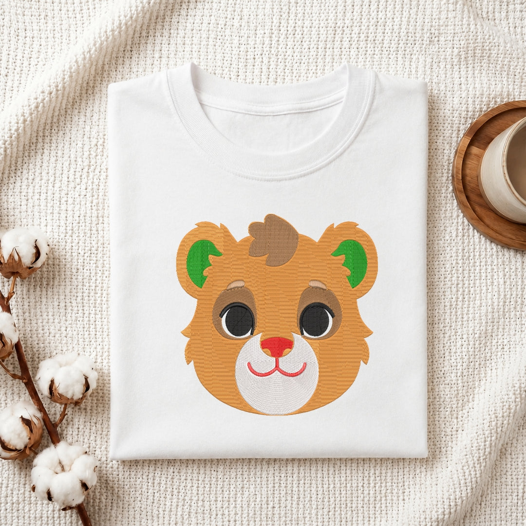 A folded white crewneck t-shirt featuring a cute cartoon bear face graphic, styled on a textured cream fabric with cotton bolls and a wooden cup in the background.