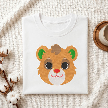 A folded white crewneck t-shirt featuring a cute cartoon bear face graphic, styled on a textured cream fabric with cotton bolls and a wooden cup in the background.