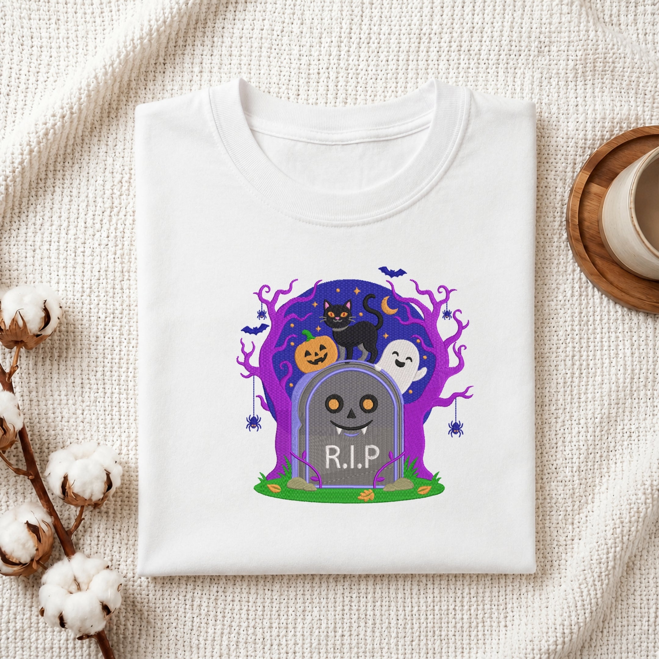 A folded white t-shirt with a colorful Halloween-themed graphic print featuring a tombstone, pumpkins, ghosts, and a purple tree, styled on a textured cream fabric with cotton bolls and a cup in the background.