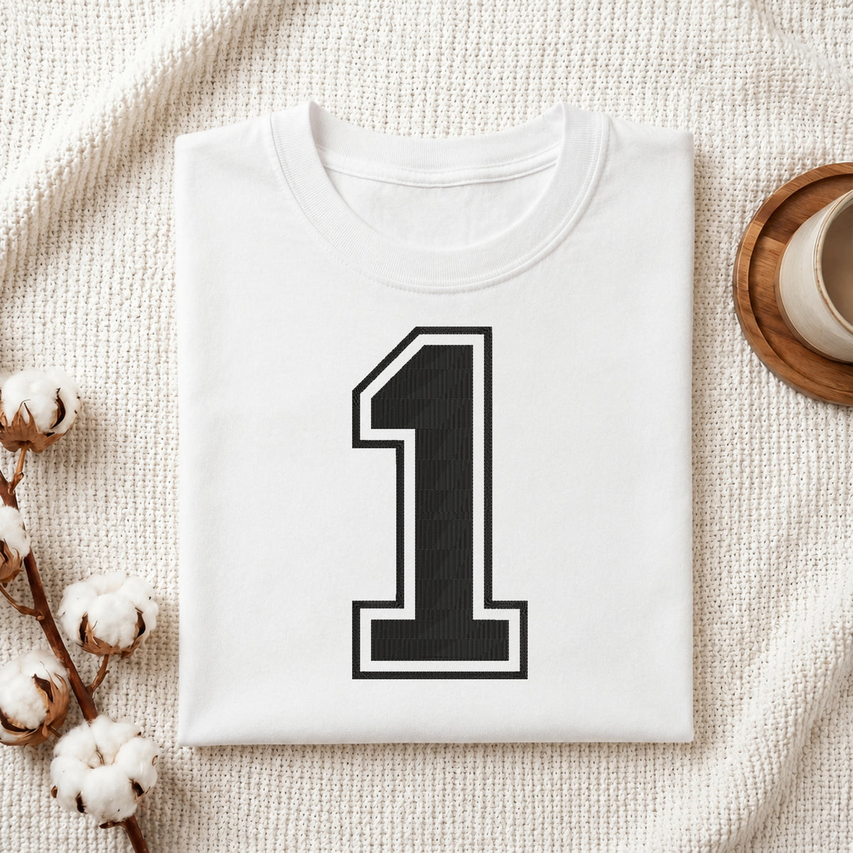 A folded white T-shirt with a large black number 1 outlined in white on the front, styled on a textured cream fabric with cotton stems and a small cup at the side.