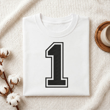A folded white T-shirt with a large black number 1 outlined in white on the front, styled on a textured cream fabric with cotton stems and a small cup at the side.