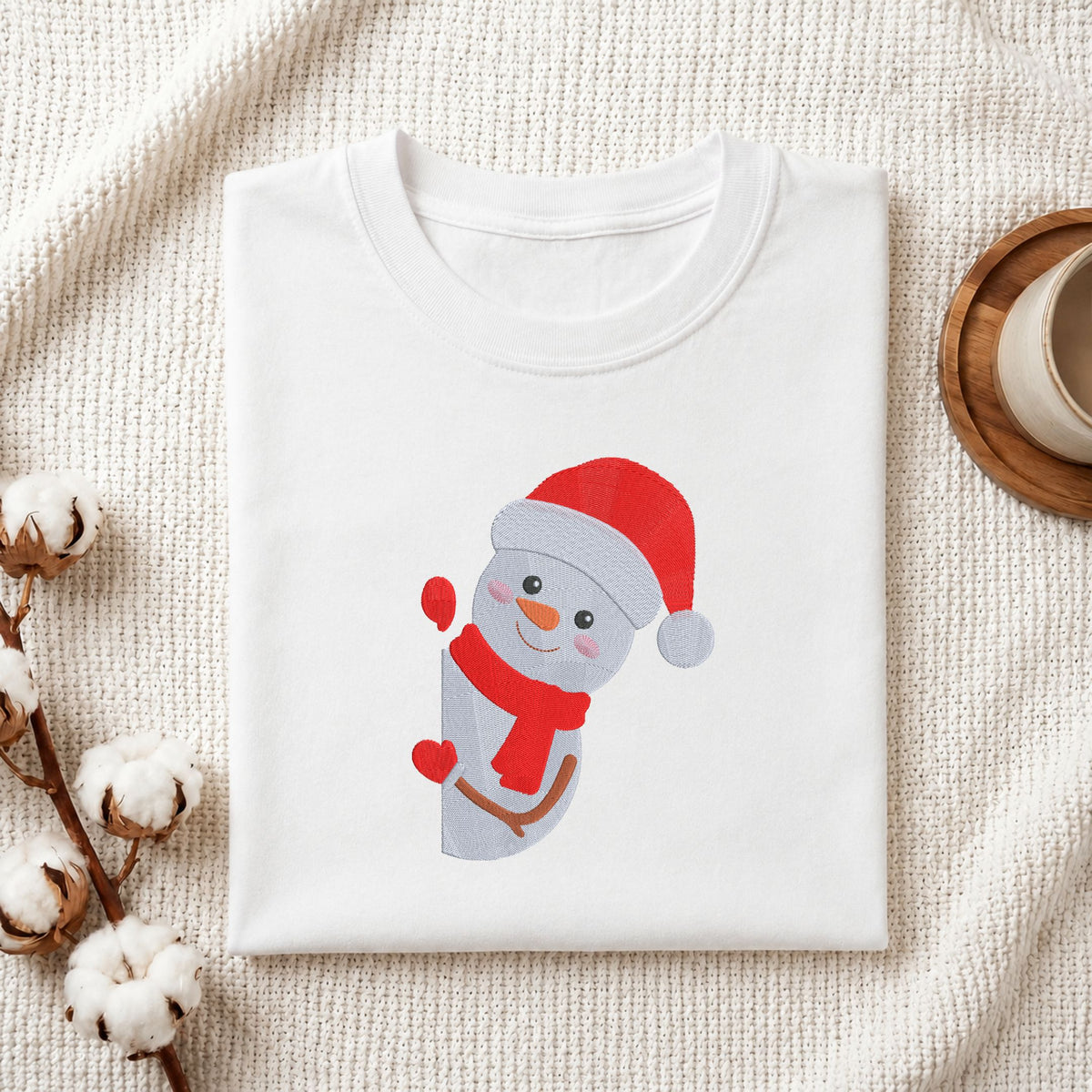 Folded white cotton t-shirt mockup featuring a cute holiday snowman machine embroidery design, styled with cotton stems and a wooden cup.