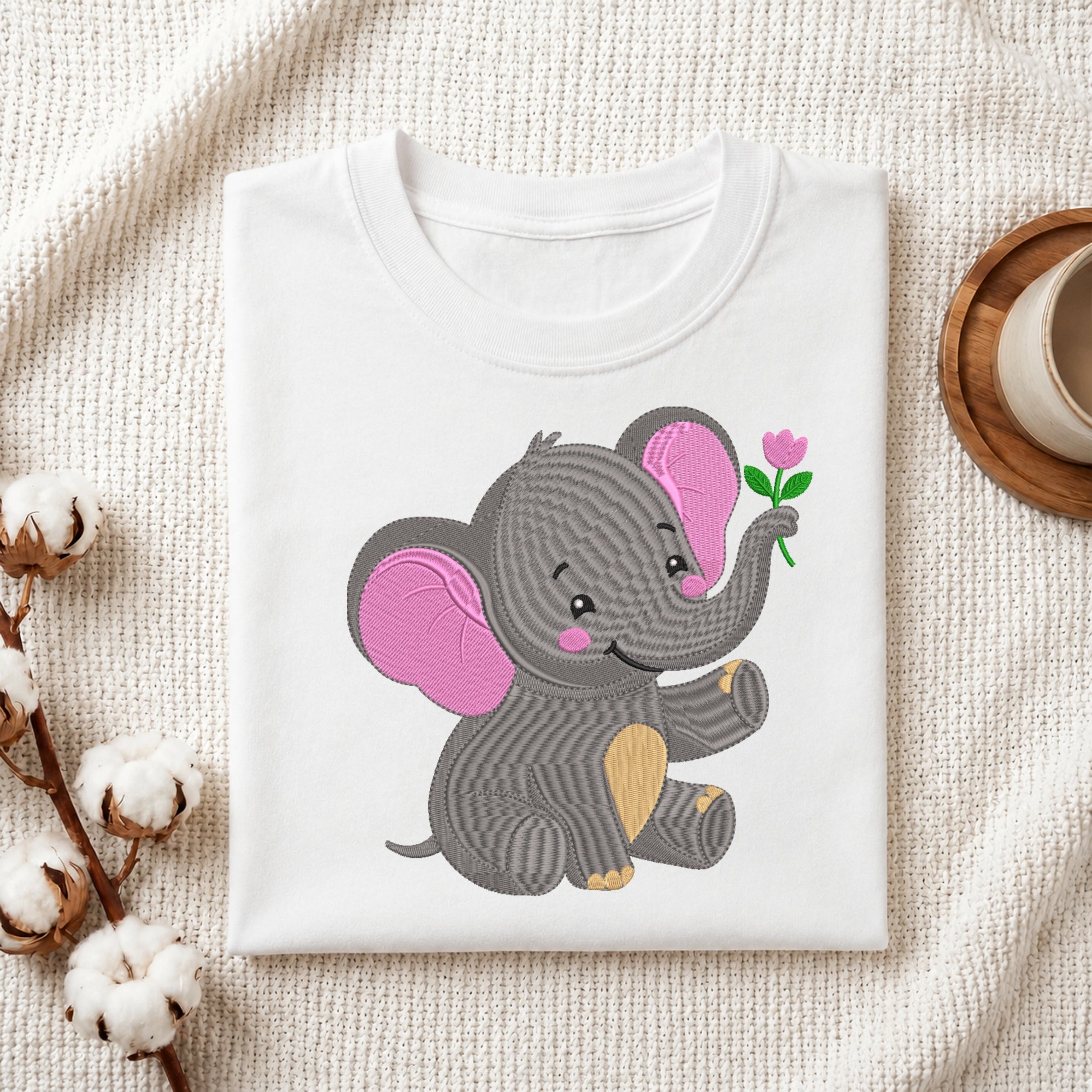 A folded white T-shirt displayed on a textured cream fabric background, featuring a cute cartoon elephant graphic in gray with pink ears and a small pink flower. Cotton stems and a cup on a coaster are styled around the shirt.