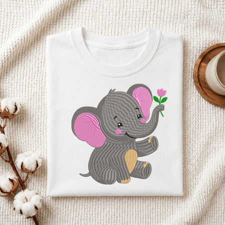 A folded white T-shirt displayed on a textured cream fabric background, featuring a cute cartoon elephant graphic in gray with pink ears and a small pink flower. Cotton stems and a cup on a coaster are styled around the shirt.