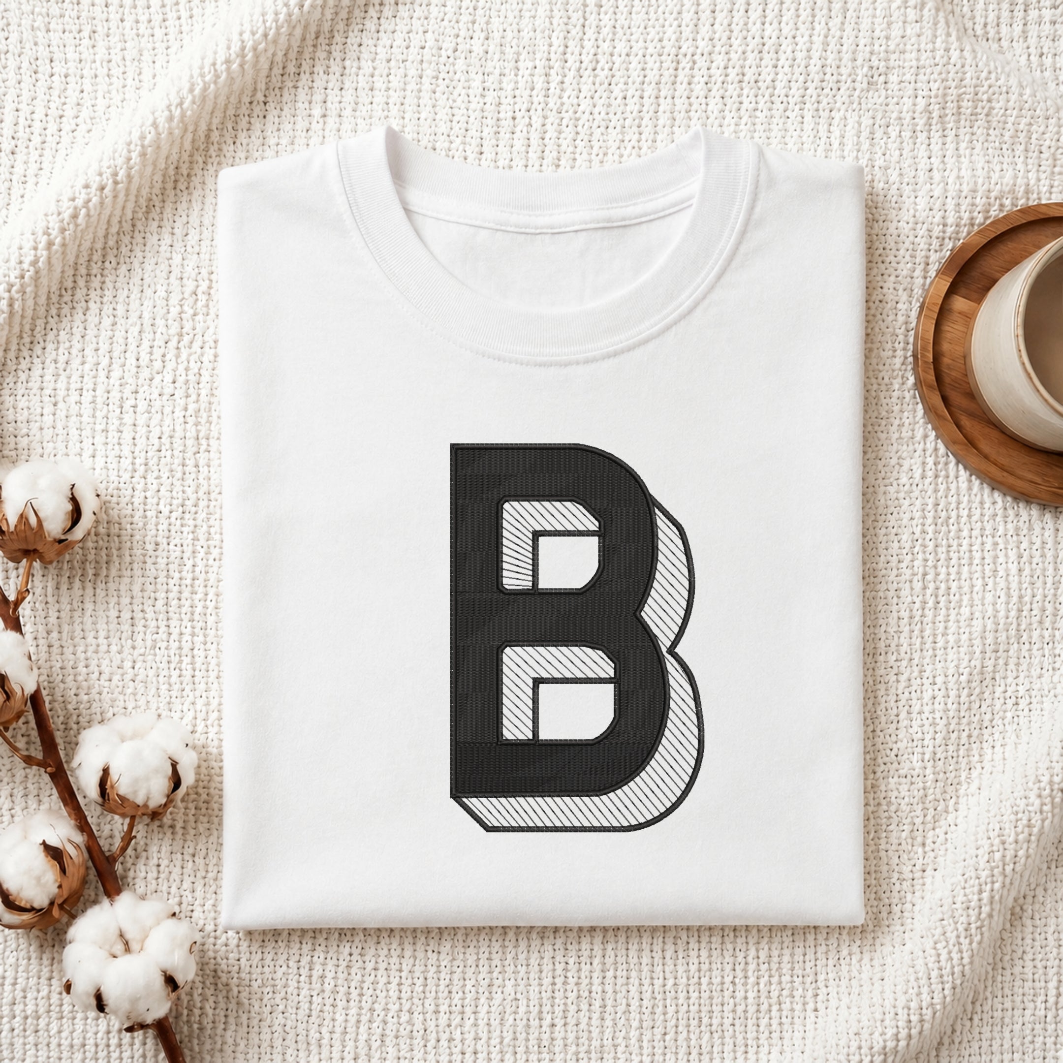 A folded white t-shirt with a large black graphic letter 'B' printed on the front, styled on a textured cream blanket with cotton stems and a cup in the background.
