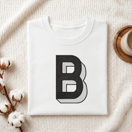 A folded white t-shirt with a large black graphic letter 'B' printed on the front, styled on a textured cream blanket with cotton stems and a cup in the background.