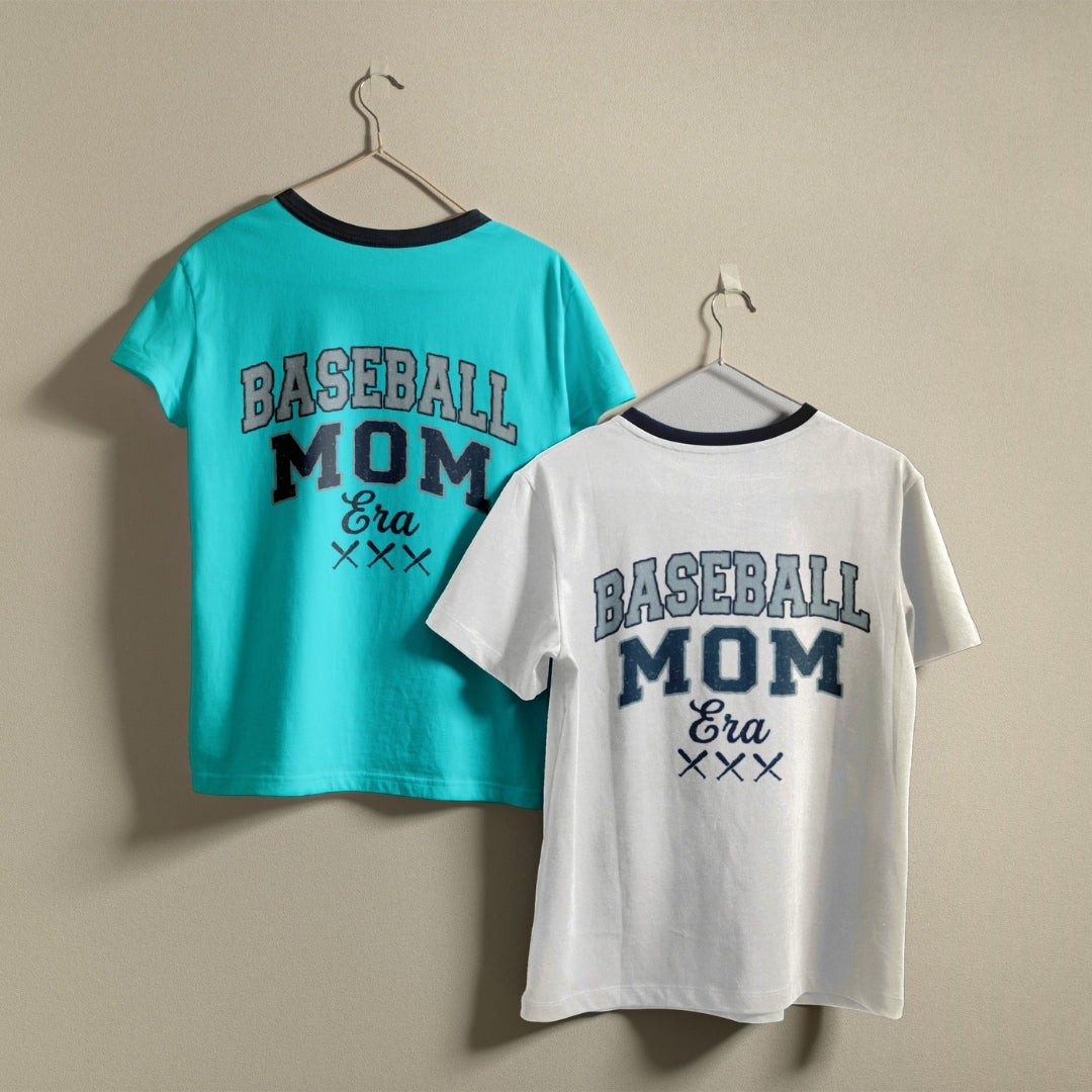 Two t-shirts hanging on a wall, one bright turquoise blue and one white, both featuring the retro collegiate "Baseball Mom Era" design.