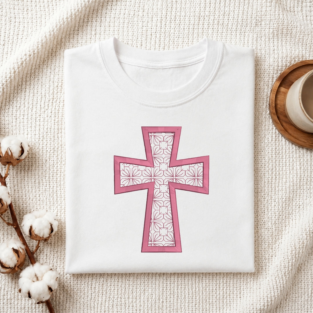 A folded white crewneck t-shirt with a pink cross graphic featuring a floral pattern, styled on a textured cream blanket with cotton bolls and a wooden cup in the background.