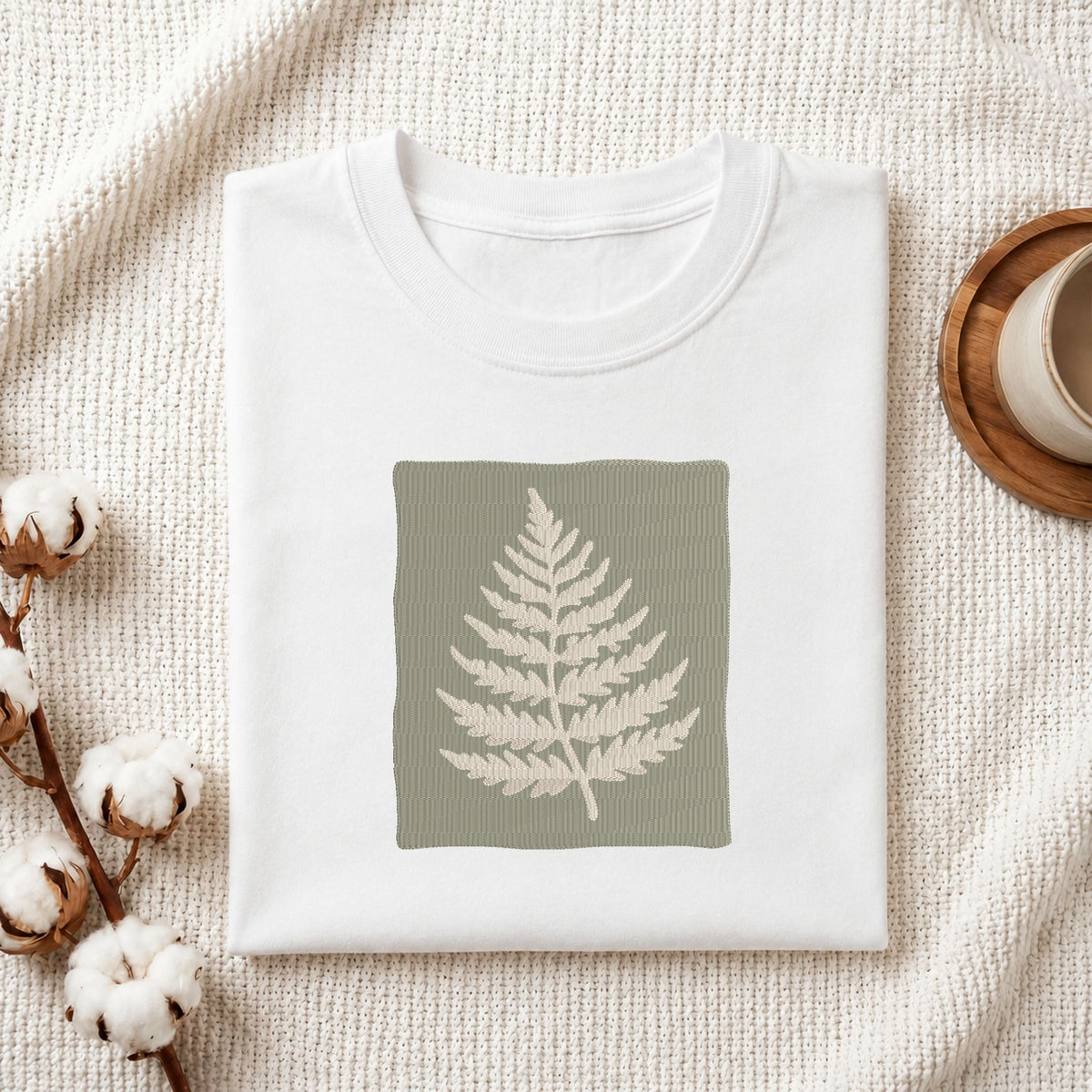 Folded white cotton t-shirt mockup featuring a sage green fern leaf machine embroidery design, styled with cotton stems and a wooden cup.