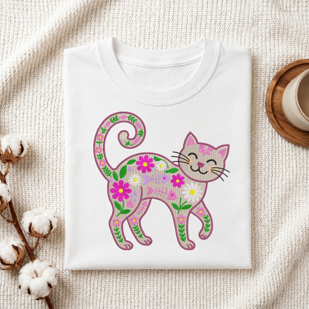 A folded white crewneck t-shirt displayed on a textured cream fabric background, featuring a colorful graphic of a smiling cat decorated with pink, purple, and green flowers and leaves.