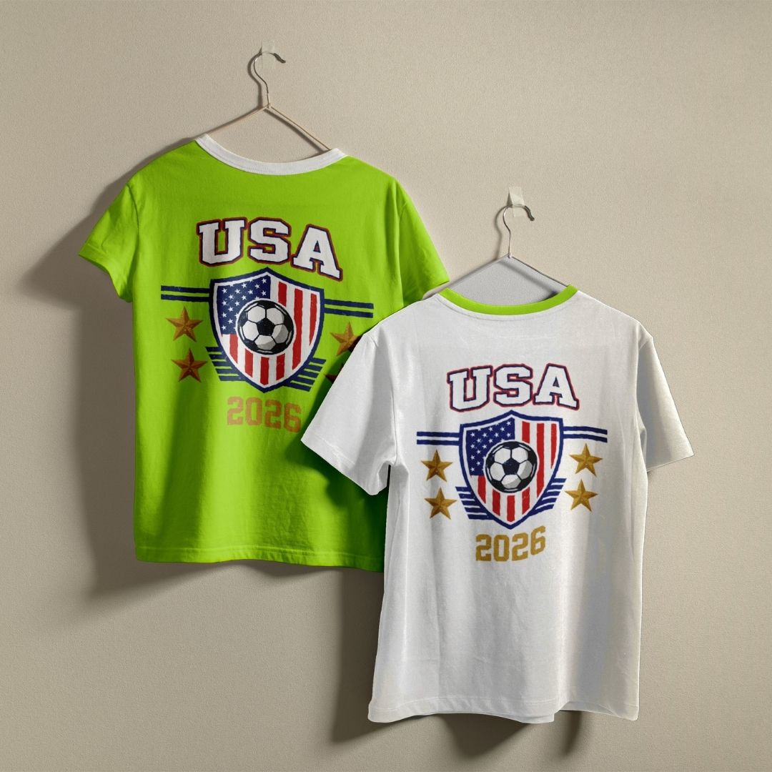 White and lime green t-shirts hanging on a wall, both displaying the vintage USA 2026 soccer shield graphic printed on the back.