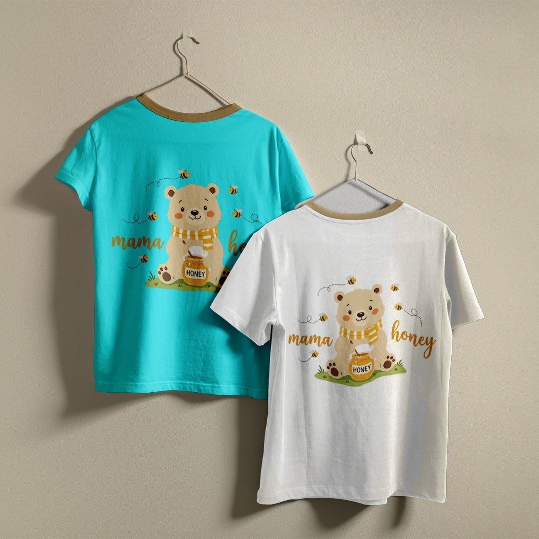 Two short-sleeve children's T-shirts hanging on hangers against a plain beige wall. One shirt is bright turquoise blue and the other is white, both featuring a cartoon bear holding a honey pot with bees and the words 'mama honey' printed on the front.