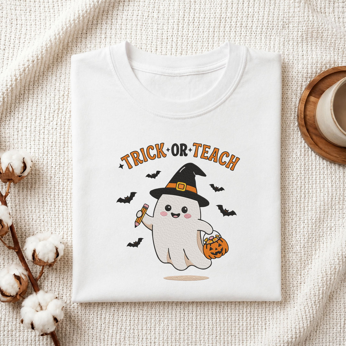Folded white cotton t-shirt mockup featuring a "Trick or Teach" teacher ghost machine embroidery design, styled with cotton stems and a wooden cup