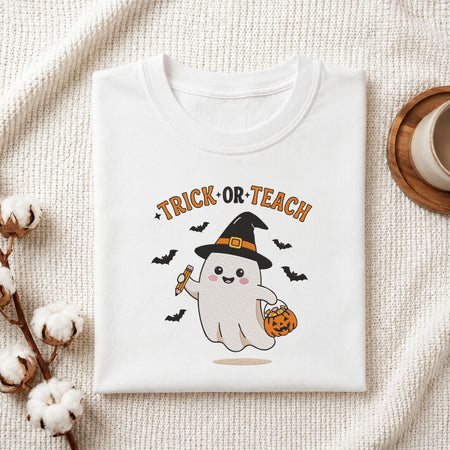 Folded white cotton t-shirt mockup featuring a "Trick or Teach" teacher ghost machine embroidery design, styled with cotton stems and a wooden cup