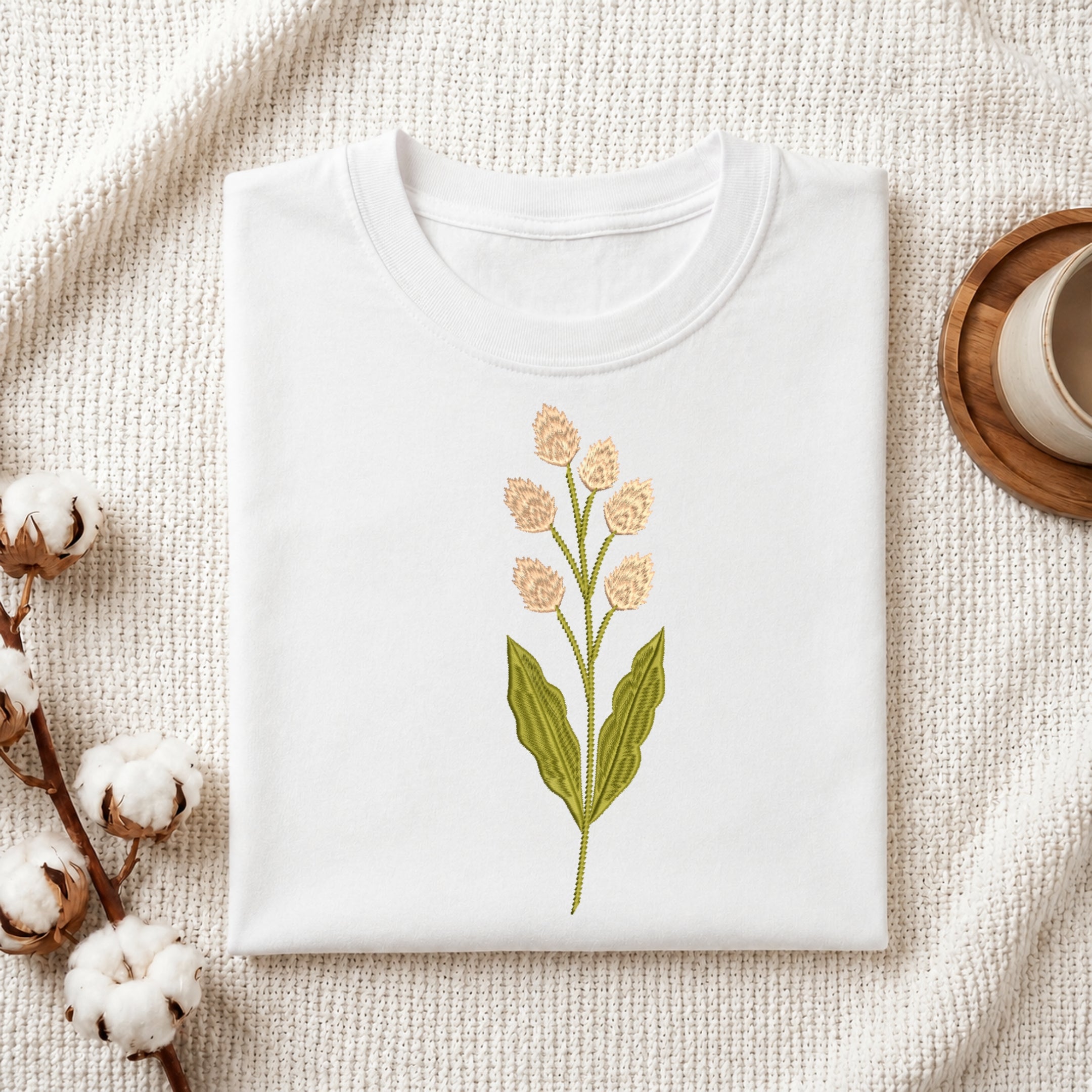 A folded white crewneck t-shirt with a central embroidered green leaf and peach-pink flower stem, styled on a textured cream knit blanket with cotton bolls and a wooden cup in the background.