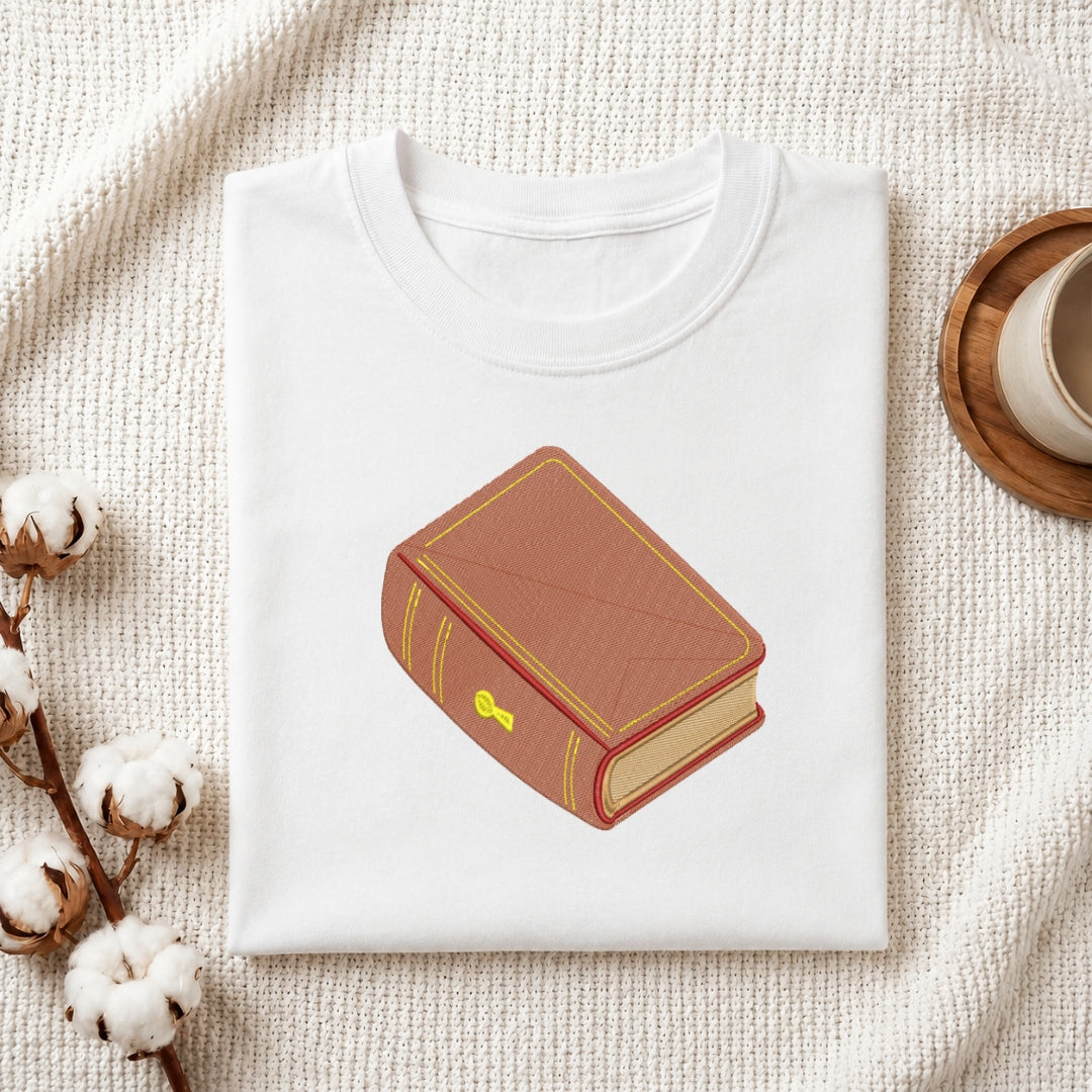 A folded white crewneck t-shirt featuring a graphic illustration of a brown leather-bound book with gold trim, styled on a textured cream knit blanket with cotton stems and a wooden cup in the background.
