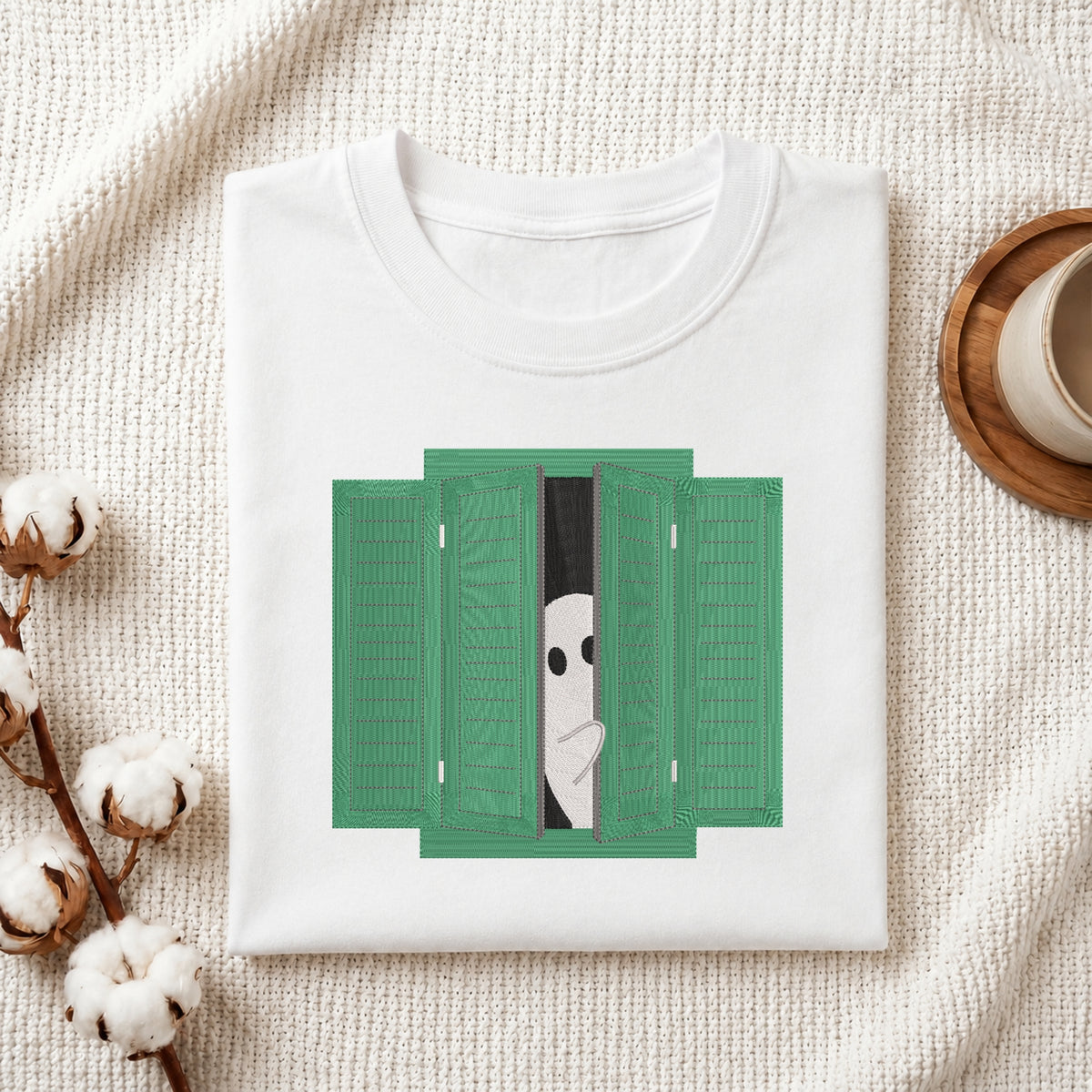 A folded white t-shirt displayed on a textured cream fabric background, decorated with cotton bolls and a wooden cup on a coaster. The t-shirt features a graphic print of a white ghost peeking out from behind green folding shutters.