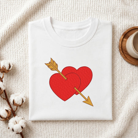A folded white crewneck t-shirt with a red heart and gold arrow graphic on the front, styled on a textured cream fabric with cotton bolls and a wooden cup.