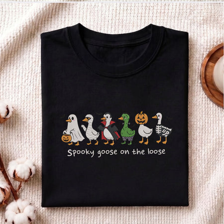  Folded black cotton t-shirt mockup featuring the "Spooky goose on the loose" Halloween costume geese machine embroidery design, styled with cotton stems and a wooden cup   