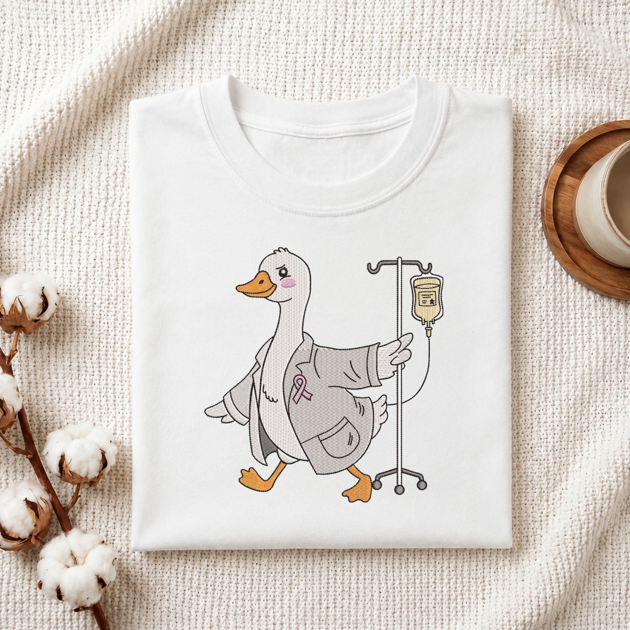 A neatly folded white crewneck t-shirt featuring a stitched cute doctor duck with IV stand design, styled with cotton branches and a wooden coaster.