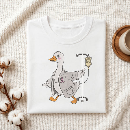 A neatly folded white crewneck t-shirt featuring a stitched cute doctor duck with IV stand design, styled with cotton branches and a wooden coaster.