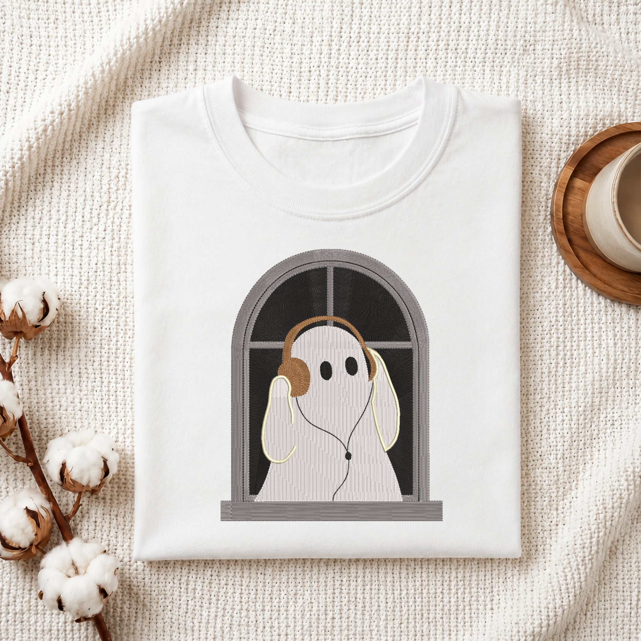 A folded white t-shirt with a graphic of a ghost wearing headphones and looking out a window, styled on a textured cream fabric with cotton bolls and a cup on a wooden coaster.