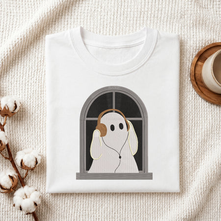 A folded white t-shirt with a graphic of a ghost wearing headphones and looking out a window, styled on a textured cream fabric with cotton bolls and a cup on a wooden coaster.